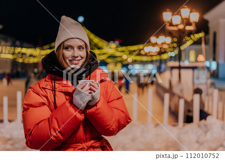 Cheerful young woman wearing warm winter clothes enjoying drinking takeaway cup of coffee outdoors on cold street at night with festive bright illumination. Pretty female walking on urban cold park Cheerful young woman wearing warm winter clothes enjoying drinking takeaway cup of coffee outdoors on cold street at night with festive bright illumination. Pretty female walking on urban cold park 112010752