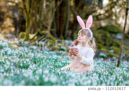 Happy little girl with Easter bunny ears eating chocolate figure in spring forest on sunny day, outdoors. Cute child with lots of snowdrop flowers. Springtime, christian holiday concept. 112012617