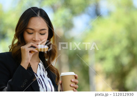 Millennial woman in business suit drinking coffee and eating sandwich in the park Millennial woman in business suit drinking coffee and eating sandwich in the park 112012930