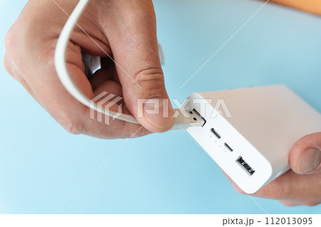 Close-up of a hand connecting a USB cable to a white power bank on a blue background. 112013095