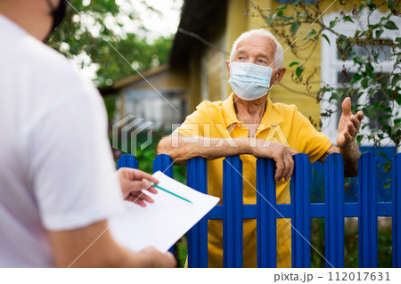 Senior man in protective mask communicating with insurance company representative while standing at fence of his country house Senior man in protective mask communicating with insurance company representative while standing at fence of his country house 112017631