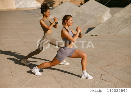 Two young sporty women in tracksuits are stretches legs outside on modern building background 112022643