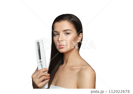 Good-looking model woman holding hair iron and straightening her healthy long dark hair isolated Good-looking model woman holding hair iron and straightening her healthy long dark hair isolated 112022678