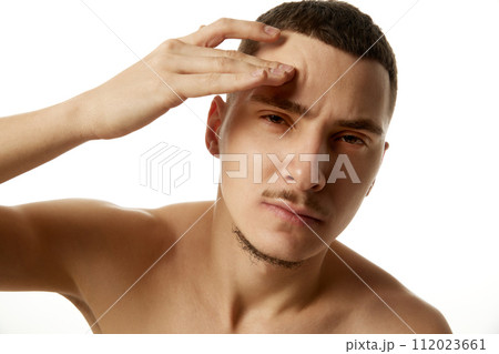 Shirtless young man holding his hand to his forehead and looking to camera with serious expression against white studio background. Shirtless young man holding his hand to his forehead and looking to camera with serious expression against white studio background. 112023661