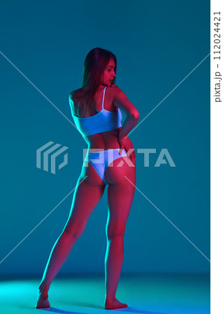 Beautiful young woman with fit, slim, tanned body posing in white underwear against blue studio background in neon light 112024421