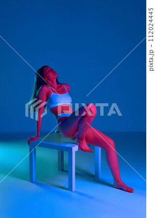 Sensual, tender, young woman with slim, fit body posing on chair in underwear against blue studio background in neon light Sensual, tender, young woman with slim, fit body posing on chair in underwear against blue studio background in neon light 112024445