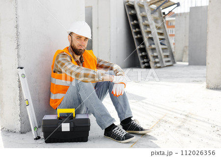 Builder repairman, foreman in safety helmet and vest sitting down to rest at workplace in building next to tool box and ruler 112026356