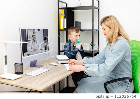 Caucasian little boy making inhalation with nebulizer at home. A young mother helping her son to inhale with a nebulizer at an online meeting with a doctor 112026754