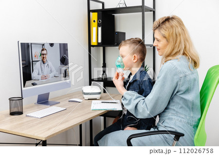 Caucasian little boy making inhalation with nebulizer at home. A young mother helping her son to inhale with a nebulizer at an online meeting with a doctor Caucasian little boy making inhalation with nebulizer at home. A young mother helping her son to inhale with a nebulizer at an online meeting with a doctor 112026758