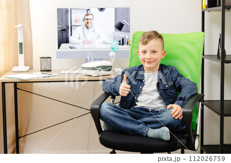 A cute smiling boy is sitting on a chair at home, and in the background is a table with a computer, at which a doctor is conducting an online consultation A cute smiling boy is sitting on a chair at home, and in the background is a table with a computer, at which a doctor is conducting an online consultation 112026759