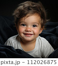 Excited baby boy on a black background smiling and looking at the camera. Studio shot of an amused small baby sitting in black blankets. Cute cheerful child with small white teeth. 112026875