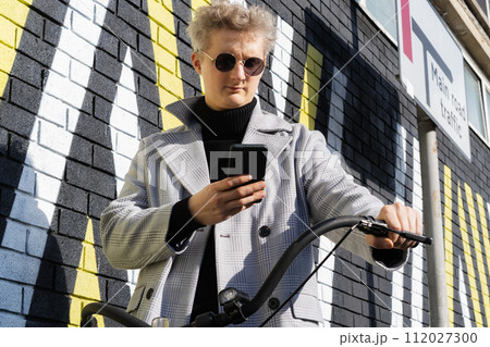 Stylish male in coat and glasses using phone while walking with retro bicycle by painted urban wall in sunny day. Neutral carbon footprint transportation. Ecofriendly mobility sustainable transport. Stylish male in coat and glasses using phone while walking with retro bicycle by painted urban wall in sunny day. Neutral carbon footprint transportation. Ecofriendly mobility sustainable transport. 112027300