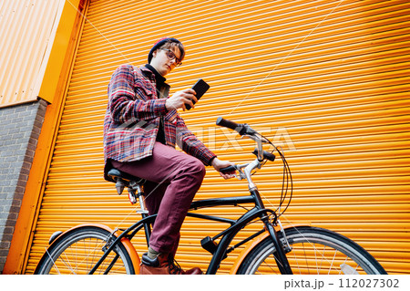 Young hipster man using mobile phone while sitting on cruiser bicycle near orange wall. Student using 5g network,rests after riding. Eco friendly transport for carbon footprint. Sustainable lifestyle Young hipster man using mobile phone while sitting on cruiser bicycle near orange wall. Student using 5g network,rests after riding. Eco friendly transport for carbon footprint. Sustainable lifestyle 112027302