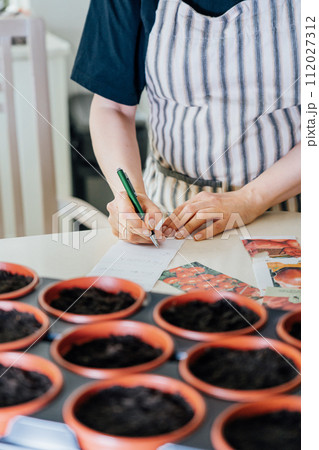 No face woman making notes on stickers for plant pots with planted seeds at home kitchen. Preparing for new kitchen garden season. Sowing seeds. Vertical card. Soft selective focus, copy space. 112027312