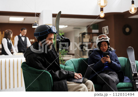 Young couple relaxing in ski resort lounge area with their digital devices enjoying winter holiday. Man with laptop and woman with cell phone taking break from skiing and snowboarding activities. 112031494