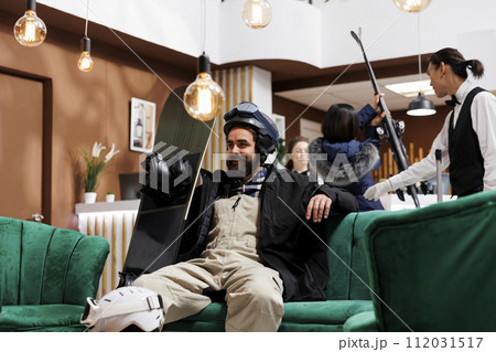 Caucasian man in hotel reception seated in cozy armchair checking his snowboarding equipment. In background bellboy helping female traveler upon arrival at front desk of luxury winter resort. 112031517
