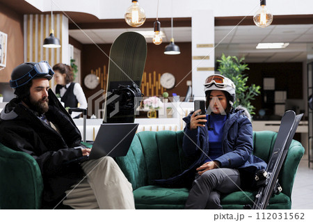 Young couple enjoys winter vacation by lounging in the ski resort lounge area with their digital gadgets. A man with a laptop and a lady with smartphone take a break from skiing and snowboarding. 112031562