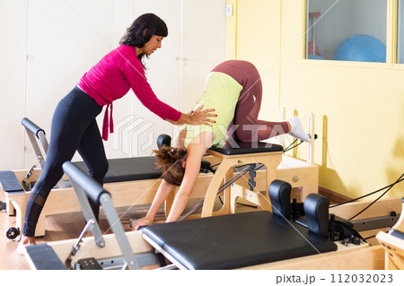 Woman practicing pilates on combo chair supervised by hispanic female trainer Woman practicing pilates on combo chair supervised by hispanic female trainer 112032023