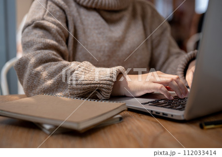 A woman in a cosy knitted sweater working remotely at a coffee shop, working on her laptop computer. A woman in a cosy knitted sweater working remotely at a coffee shop, working on her laptop computer. 112033414