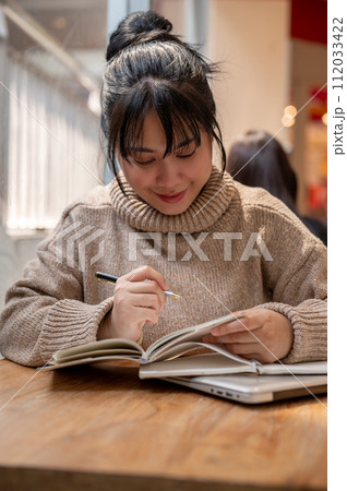 A charming Asian woman is listing her ideas in a book, working remotely at a coffee shop. 112033422
