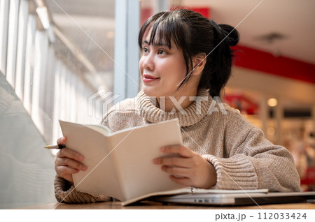 A woman is looking out the window and daydreaming while reading a book in a coffee shop. 112033424