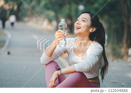 Sport fitness concept, A joyful young woman takes a water break outdoors after exercising, wearing earphones and sportswear, embodying a healthy lifestyle. Sport fitness concept, A joyful young woman takes a water break outdoors after exercising, wearing earphones and sportswear, embodying a healthy lifestyle. 112034888