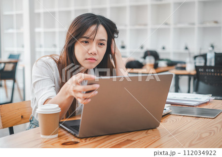 A young woman looks frustrated and stressed while concentrating on her laptop screen in a bustling cafe environment. 112034922