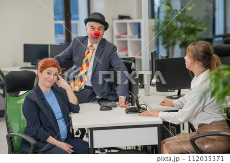 An elderly Caucasian man in a clown costume amuses two Caucasian women in the office.  112035371