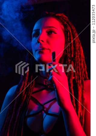 Caucasian girl with dreadlocks smokes a vape in red blue light.  112035473