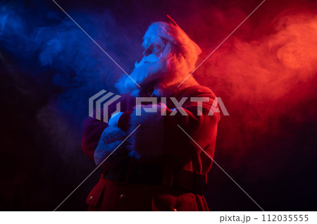 Santa Claus stands with his arms crossed on his chest in blue red neon light and smoke.  112035555