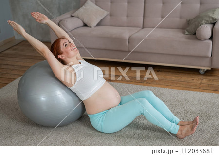 Pregnant red-haired woman doing exercises on fitness ball at home.  112035681
