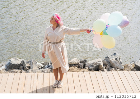 A woman with colored hair and an armful of balloons walks along the river embankment. 112035712