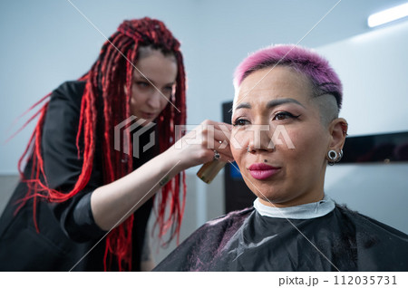 The hairdresser shaves the temple of a female client. Rear view of a woman with short pink hair in a barbershop. 112035731