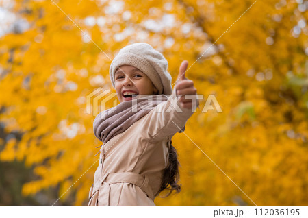Happy caucasian girl in a beige coat and beret walks in the park in autumn. Schoolgirl showing thumbs up. Happy caucasian girl in a beige coat and beret walks in the park in autumn. Schoolgirl showing thumbs up. 112036195