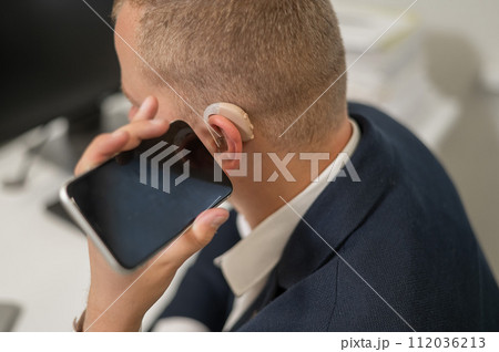 Caucasian man with a hearing aid talking on the phone on speakerphone.  112036213