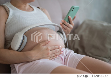 A pregnant woman turns on music for her child on a smartphone. Headphones on the tummy. A pregnant woman turns on music for her child on a smartphone. Headphones on the tummy. 112036242