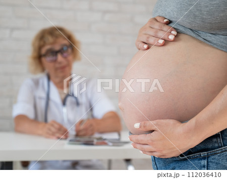 Doctor obstetrician gynecologist at his desk in the background. Close-up of a pregnant woman's belly. Doctor obstetrician gynecologist at his desk in the background. Close-up of a pregnant woman's belly. 112036410