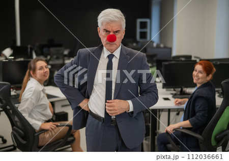 Two Caucasian women look at a serious elderly man with a clown nose in the office. 112036661