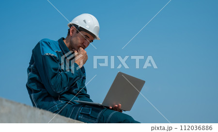 Caucasian male builder in hardhat sits on floor slabs and uses laptop at construction site.  112036886