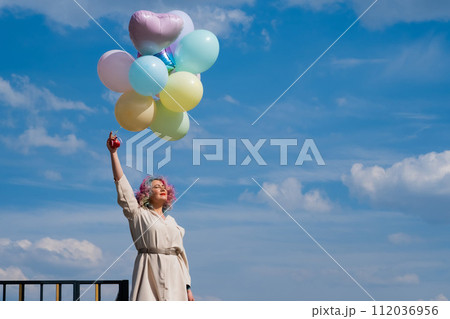 A woman with colored hair with an armful of balloons against a blue sky. 112036956