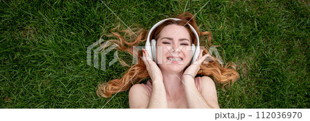Top view of a young red-haired woman lying on the grass and listening to music on headphones. Widescreen.  112036970