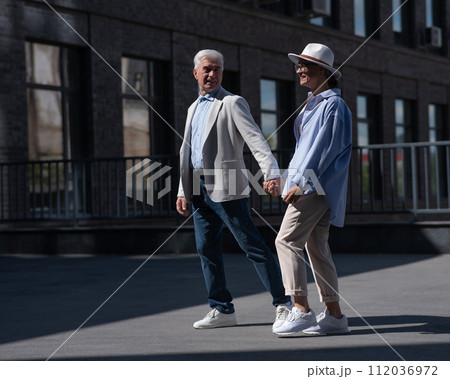Stylish mature couple. Gray-haired man and woman holding hands while walking around the city. Stylish mature couple. Gray-haired man and woman holding hands while walking around the city. 112036972