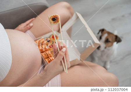 A pregnant woman sits on the sofa and eats rolls. Jack Russell Terrier dog sits on the floor and begs for food from its owner.  112037004