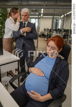 Elderly Caucasian man and woman gossiping behind their sleeping pregnant colleague in the office. Vertical photo. Elderly Caucasian man and woman gossiping behind their sleeping pregnant colleague in the office. Vertical photo. 112037033