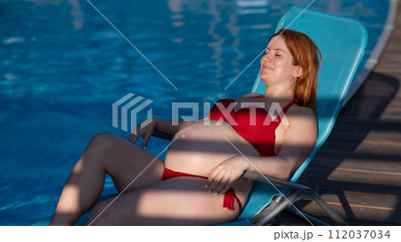Red-haired pregnant woman in a red bikini sunbathes lying on a sun lounger next to the pool. Red-haired pregnant woman in a red bikini sunbathes lying on a sun lounger next to the pool. 112037034