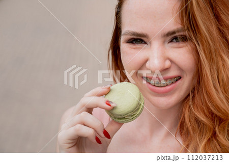 Young red-haired woman with braces eating macaron cake. Young red-haired woman with braces eating macaron cake. 112037213