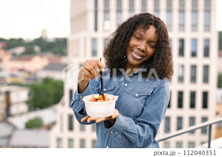 African businesswoman eating healthy salad standing on rooftop on a city view. African businesswoman eating healthy salad standing on rooftop on a city view. 112040351