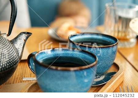 Brewing and Serving Tea. Teapot Pouring Green Tea in Blue Ceramic Cup on Wooden Cafe Table. Two Pottery Handmade Mugs Early in the Morning. Trendy Authentic Porcelain Set. Breakfast Concept. Tea Time Brewing and Serving Tea. Teapot Pouring Green Tea in Blue Ceramic Cup on Wooden Cafe Table. Two Pottery Handmade Mugs Early in the Morning. Trendy Authentic Porcelain Set. Breakfast Concept. Tea Time 112045232
