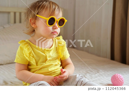 Child playing with colorful plastic toys on beige cozy bed in sunny white bedroom. Educational game for baby and toddler. Young girl play indoors. Wearing sunglasses. Kid preparing to vacation, travel Child playing with colorful plastic toys on beige cozy bed in sunny white bedroom. Educational game for baby and toddler. Young girl play indoors. Wearing sunglasses. Kid preparing to vacation, travel 112045366