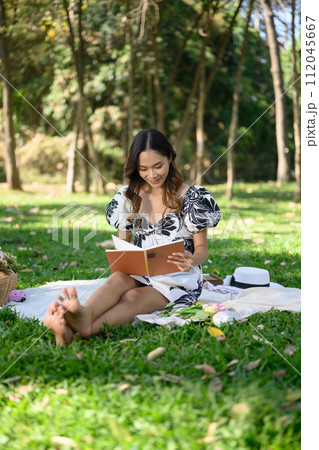 Full length of happy young woman sitting on grass with barefoot and reading book 112045667